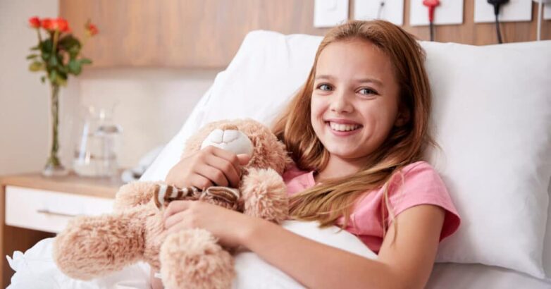 Portrait Of Girl Lying In Bed In Hospital Ward Hugging Teddy Bear