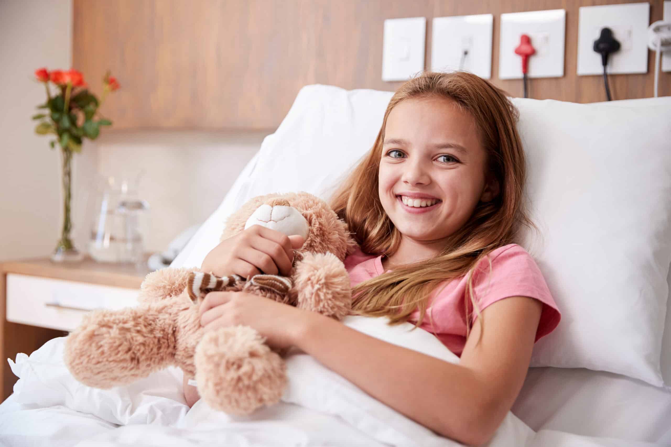 Portrait Of Girl Lying In Bed In Hospital Ward Hugging Teddy Bear